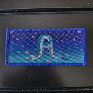 May include: A rectangular blue patch with a white and blue dragon design. The background fades from dark to light blue, with pink petal shapes. The patch has a blue border. The design is reminiscent of Japanese art.