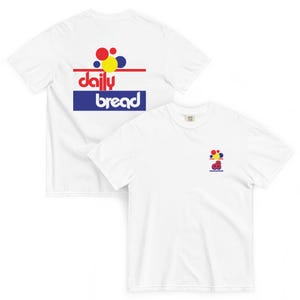 Könnte beinhalten: Ein weißes kurzärmeliges T-Shirt mit einer Grafik auf der Rückseite und einer kleineren Version auf der Vorderseite. Die Grafik zeigt rote, gelbe und blaue Kreise über einer roten Linie und dem Text daily bread in Blau auf blauem Hintergrund.