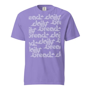 Puede incluir: Una camiseta de manga corta de color morado claro con un estampado gráfico repetitivo en color lavanda pálido. El texto daily bread aparece en una fuente estilizada, ondulada y similar a una nube que cubre el centro del pecho de la prenda.