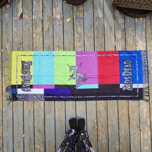 May include: A rectangular scarf with a colorful, geometric design. The scarf features a vibrant mix of colors, including yellow, blue, green, purple, red, and black. The words "Zeds Dead" are printed on the scarf. The scarf has fringe on the ends.