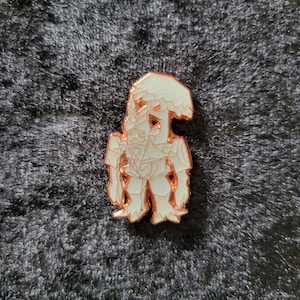 Pode incluir: Um alfinete de esmalte branco e cobre em forma de uma figura estilizada semelhante a um cogumelo. O alfinete tem um design geométrico com uma aparência texturizada. A figura está em pé com um chapéu de cogumelo para a cabeça.