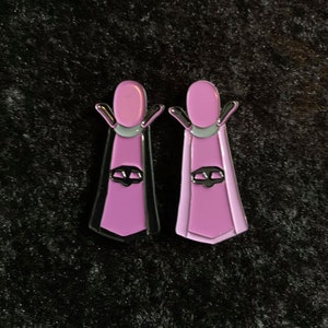 May include: Two pink enamel pins with black accents. Each pin features a stylized figure with a skull and crossbones design.