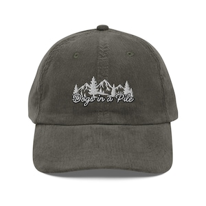 May include: Olive green corduroy baseball cap with a curved brim. The cap features white embroidered text and a mountain and tree design that reads "Dogs in a Pile".