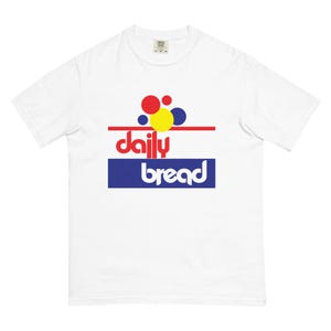 Può includere: Una t-shirt bianca a maniche corte con un design grafico sul petto. Il motivo include cerchi rossi, gialli e blu sopra una linea orizzontale rossa, con il testo rosso daily e il testo bianco bread all'interno di un rettangolo blu sottostante.