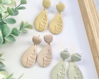 May include: Three pairs of dangle earrings with a teardrop shape and a leaf design. The earrings are made of polymer clay and come in three colors: beige, light brown, and light green.