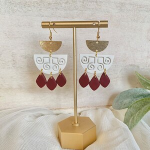 May include: A pair of handmade earrings displayed on a gold stand. The earrings feature a gold half-circle top, a white, semi-circular middle section with a swirl design, and three maroon teardrop-shaped dangles. The earrings are designed for pierced ears.