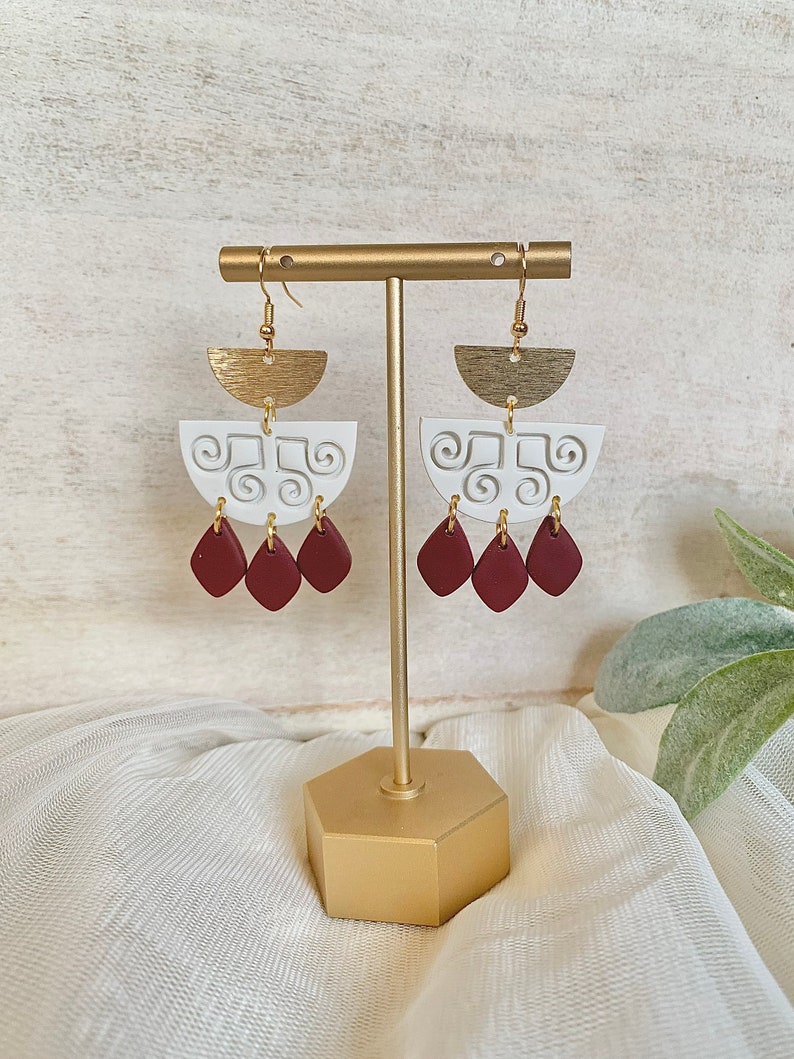 May include: A pair of handmade earrings displayed on a gold stand. The earrings feature a gold half-circle top, a white, semi-circular middle section with a swirl design, and three maroon teardrop-shaped dangles. The earrings are designed for pierced ears.