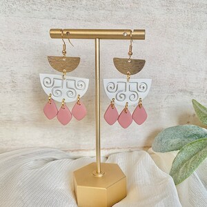 May include: A pair of handmade earrings displayed on a gold stand. The earrings feature a brass half-circle top, a white, semi-circular middle section with a swirl design, and three pink teardrop-shaped dangles. The earrings are designed for pierced ears.