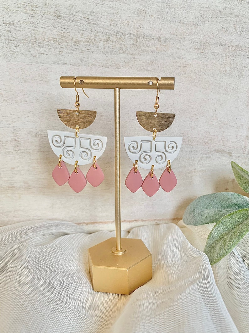 May include: A pair of handmade earrings displayed on a gold stand. The earrings feature a brass half-circle top, a white, semi-circular middle section with a swirl design, and three pink teardrop-shaped dangles. The earrings are designed for pierced ears.