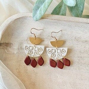 May include: A pair of handmade earrings with a unique design. The earrings feature gold-colored half-circle tops, white clay mid-sections with a swirl pattern, and three maroon-colored teardrop shapes hanging below. The earrings are designed to be worn with a hook.
