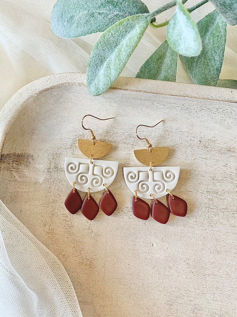May include: A pair of handmade earrings with a unique design. The earrings feature gold-colored half-circle tops, white clay mid-sections with a swirl pattern, and three maroon-colored teardrop shapes hanging below. The earrings are designed to be worn with a hook.