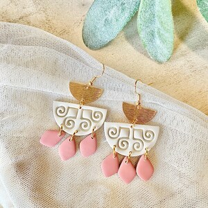 May include: A pair of handmade earrings with a gold-tone half-circle top, white center with a swirl design, and pink teardrop-shaped dangles. The earrings hang from gold-colored ear wires. The background is a neutral color with a sheer fabric.