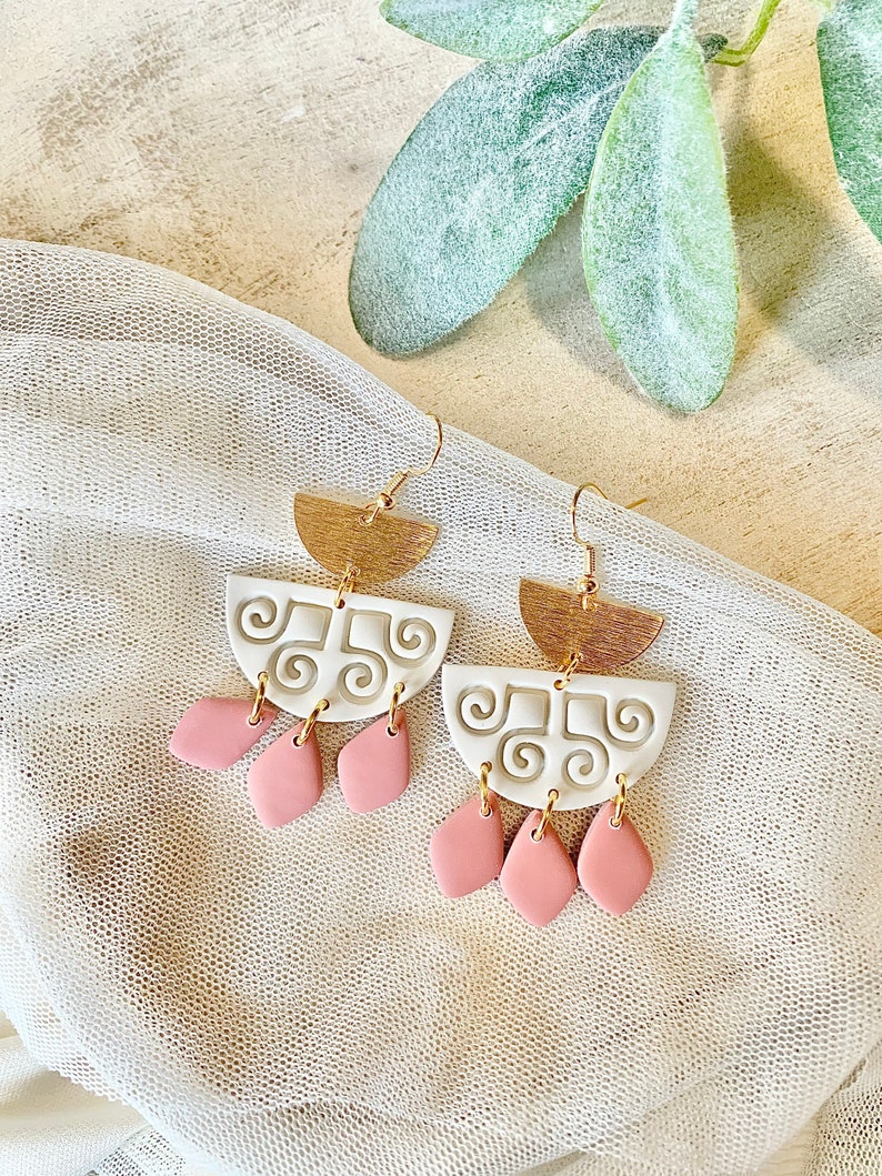 May include: A pair of handmade earrings with a gold-tone half-circle top, white center with a swirl design, and pink teardrop-shaped dangles. The earrings hang from gold-colored ear wires. The background is a neutral color with a sheer fabric.