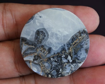 Puede incluir: Un cabujón redondo de ágata de color blanco, gris y dorado con un patrón dendrítico. La ágata tiene una superficie lisa y pulida.