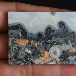 May include: A close-up of a polished, gray, white, and orange agate cabochon with a swirling pattern. The cabochon has a smooth, polished surface and is set against a dark background.