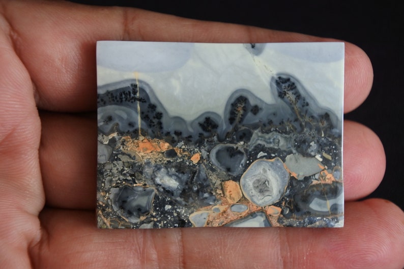 May include: A close-up of a polished, gray, white, and orange agate cabochon with a swirling pattern. The cabochon has a smooth, polished surface and is set against a dark background.