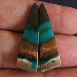 May include: A pair of triangular shaped cabochons with a layered pattern of brown, white, blue and green colors. The cabochons are cut from a natural stone.