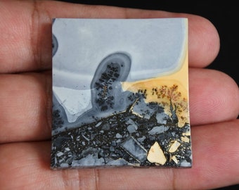 May include: A square, polished stone with a unique pattern. The stone features a blend of gray, black, and tan hues, with intricate veining and patterns resembling a landscape. The stone is held in a hand, showcasing its natural beauty.