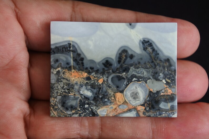 May include: A close-up of a polished, gray, black, and orange agate slab with a unique, swirling pattern. The slab has a smooth, polished surface and is approximately 2 inches by 3 inches.