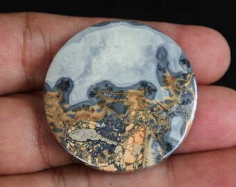 Puede incluir: Un cabujón de ágata redondo y pulido con un patrón único de gris, blanco, marrón y negro. La piedra tiene un aspecto natural y orgánico.