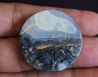 Puede incluir: Una piedra pulida y redonda con un patrón único. La mitad superior presenta un diseño en capas en gris claro y blanco, mientras que la mitad inferior es una mezcla texturizada más oscura de negro y gris. Finas vetas de color amarillo dorado atraviesan la piedra.