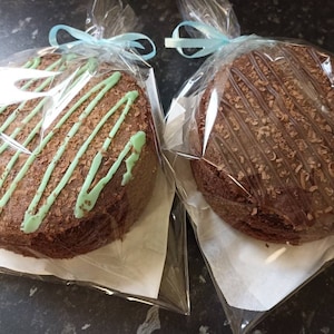 May include: Two chocolate cakes, round in shape, presented in clear cellophane with light blue ribbon ties. One is decorated with green icing and chocolate flakes, the other with chocolate icing and flakes.