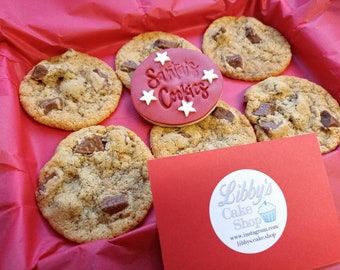 May include: A selection of chocolate chip cookies with chocolate chunks, presented in a red box. One cookie is decorated with the words "Santa's Cookies" and star shapes. A red card with a white Libby's Cake Shop logo is also visible.
