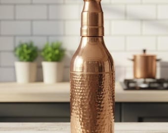 May include: A copper-colored bottle with a textured, hammered design. The bottle has a narrow neck and a wider base, with a shiny, metallic finish. The kitchen setting includes a white tile backsplash and wooden shelves.
