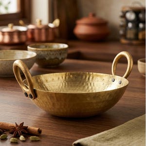 May include: A hammered brass bowl with two loop handles. The bowl has a textured surface and is sitting on a wooden table. Other kitchenware and spices are in the background.