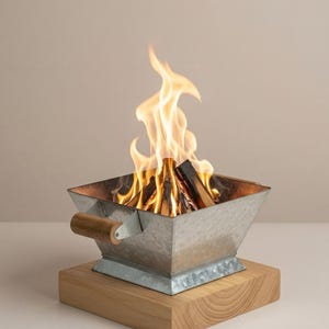 May include: A small, galvanised metal fire pit with a wooden handle and base. Flames rise from burning wood inside the square container. The fire pit is a decorative item for outdoor or indoor use.
