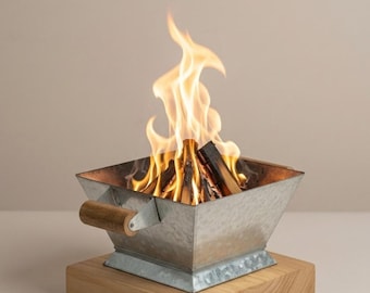 May include: A small, galvanized metal fire pit with a wooden handle and base. Flames rise from burning wood inside the square container. The fire pit is a decorative item for outdoor or indoor use.
