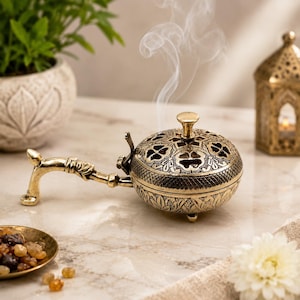 May include: A round, gold-toned metal incense burner with an ornate, perforated lid and a long, decorative handle. The burner features intricate engraved patterns and rests on small feet, with a thin wisp of white smoke rising from the top.