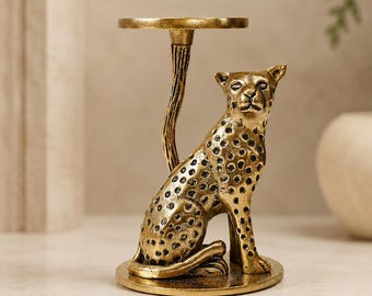 Puede incluir: Un portavelas de metal en tono dorado con la figura de un leopardo sentado con manchas negras. El leopardo se asienta sobre una base circular y sostiene una plataforma redonda y plana sobre su cabeza con un tallo delgado y texturizado.