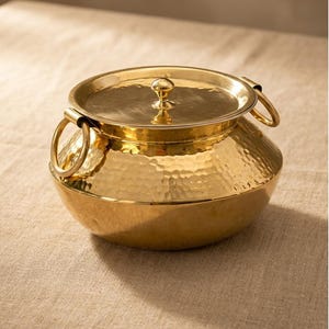 Puede incluir: Una olla de metal dorado martillado con tapa y dos asas de anillo. La olla tiene una base redondeada y una tapa con un pequeño pomo. La superficie de la olla tiene un aspecto texturizado y con hoyuelos. La olla está sobre una superficie de color neutro.