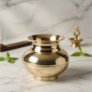 May include: A gold-colored, round brass pot with horizontal ridges. The pot has a wide mouth and a small base. Incense and a small oil lamp are in the background. The pot is on a white marble surface.