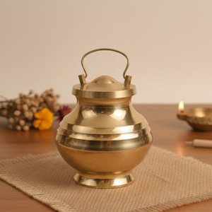 May include: A golden-colored metal pot with a lid and handle. The pot has horizontal bands around its body and sits on a small base. It is placed on a tan fabric, with a blurred background featuring flowers and a candle.