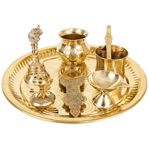 May include: A brass puja thali set featuring a bell, a diya, a kalash, a spoon, and a small incense burner. The set is arranged on a round brass tray with a decorative pattern.
