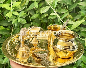 Puede incluir: Una bandeja de metal dorado con un diseño decorativo, que contiene un conjunto de pequeños objetos ceremoniales. Los objetos incluyen una campana, una cuchara, una taza pequeña, un diya y una olla pequeña.
