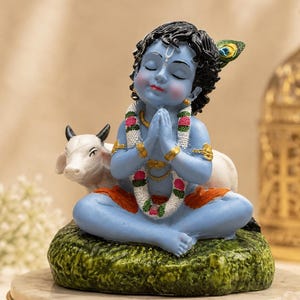 May include: A blue-skinned figurine of a child deity sitting in a cross-legged position with hands pressed together in prayer. The figure wears a white floral garland with pink accents, orange lower garments, and a peacock feather in black hair.