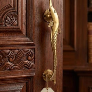 May include: A gold-colored dolphin-shaped door pull mounted on a dark wooden door. The door pull features a detailed design of a dolphin with a textured body and a curved handle. A tag with the text "Dolphin Door Pull" hangs from the handle.