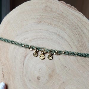 Puede incluir: Un collar choker de macram&eacute; verde con tres colgantes dorados en forma de sol. El collar est&aacute; adornado con peque&ntilde;as cuentas doradas y se exhibe sobre una superficie de madera. Esta joya hecha a mano es un accesorio bohemio.