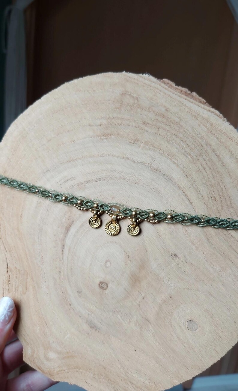 Puede incluir: Un collar choker de macram&eacute; verde con tres colgantes dorados en forma de sol. El collar est&aacute; adornado con peque&ntilde;as cuentas doradas y se exhibe sobre una superficie de madera. Esta joya hecha a mano es un accesorio bohemio.