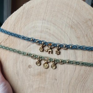 Puede incluir: Dos gargantillas de macram&eacute; con abalorios dorados. Una es azul, la otra verde. Los abalorios incluyen peque&ntilde;as flores, soles y un elefante. Las gargantillas se exhiben sobre una superficie de madera.