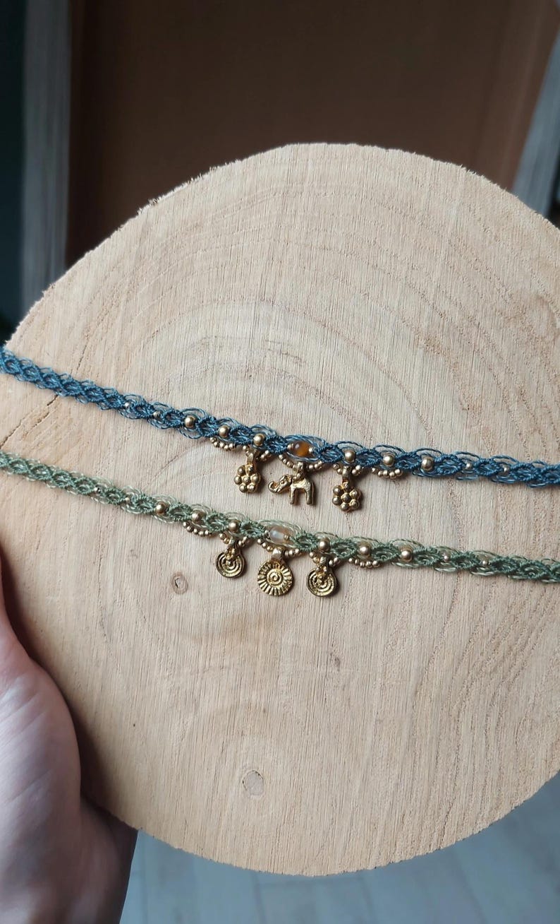 Puede incluir: Dos gargantillas de macram&eacute; con abalorios dorados. Una es azul, la otra verde. Los abalorios incluyen peque&ntilde;as flores, soles y un elefante. Las gargantillas se exhiben sobre una superficie de madera.