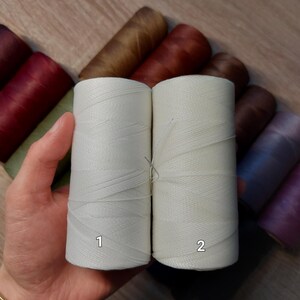 Puede incluir: Dos carretes de hilo blanco roto se sostienen en una mano, etiquetados 1 y 2. Varios otros carretes de hilo en tonos marr&oacute;n, burdeos y morado son visibles en el fondo. El hilo parece estar bien enrollado y listo para usar.