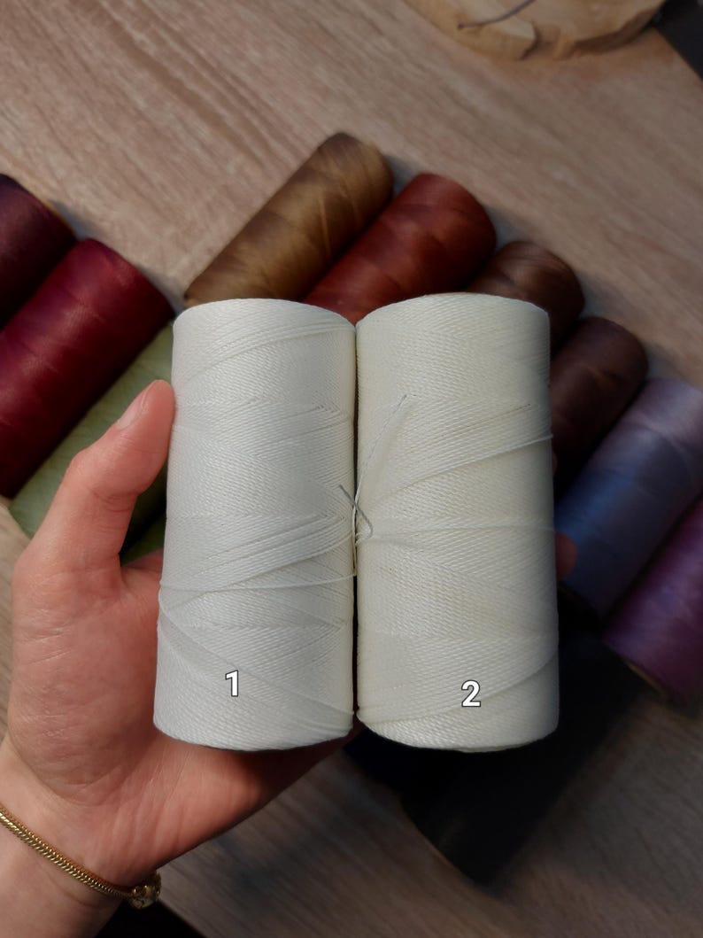 Puede incluir: Dos carretes de hilo blanco roto se sostienen en una mano, etiquetados 1 y 2. Varios otros carretes de hilo en tonos marr&oacute;n, burdeos y morado son visibles en el fondo. El hilo parece estar bien enrollado y listo para usar.