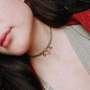 Puede incluir: Primer plano de un collar con un cord&oacute;n trenzado gris verdoso y peque&ntilde;os abalorios dorados. El collar se lleva en el cuello de una persona, con un su&eacute;ter rojo visible en el fondo. Tambi&eacute;n se ven el pelo oscuro y la piel de la persona.