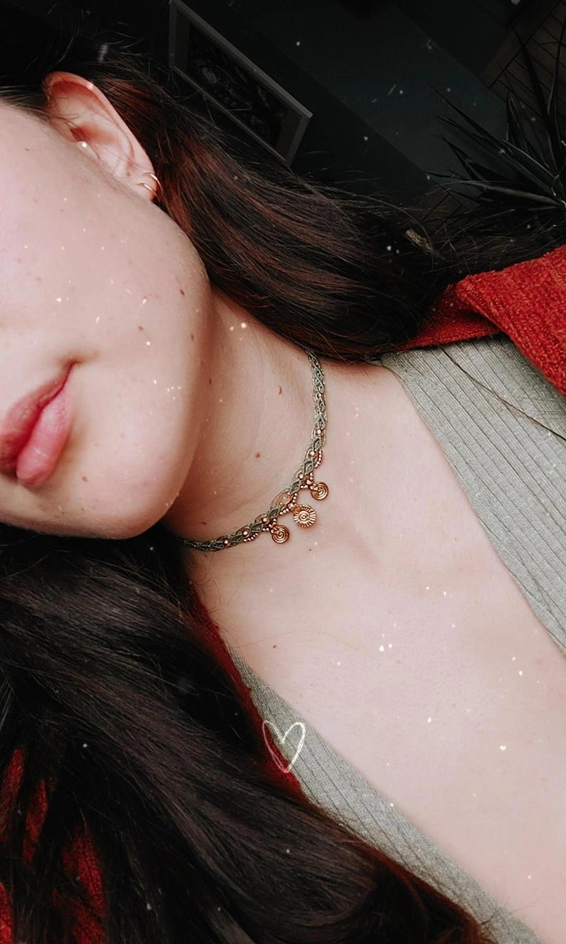 Puede incluir: Primer plano de un collar con un cord&oacute;n trenzado gris verdoso y peque&ntilde;os abalorios dorados. El collar se lleva en el cuello de una persona, con un su&eacute;ter rojo visible en el fondo. Tambi&eacute;n se ven el pelo oscuro y la piel de la persona.