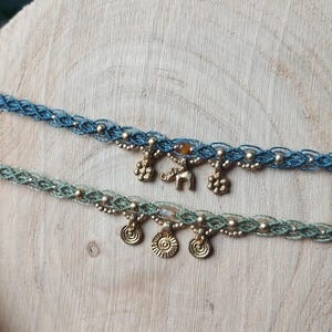 Puede incluir: Dos gargantillas de macram&eacute; hechas a mano. Una es azul con cuentas y abalorios dorados, incluido un elefante. La otra es verde con cuentas doradas y abalorios en forma de sol. Ambas se exhiben sobre una superficie de madera.