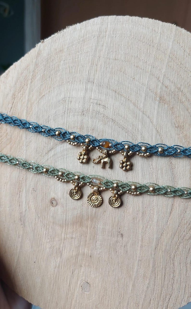 Puede incluir: Dos gargantillas de macram&eacute; hechas a mano. Una es azul con cuentas y abalorios dorados, incluido un elefante. La otra es verde con cuentas doradas y abalorios en forma de sol. Ambas se exhiben sobre una superficie de madera.